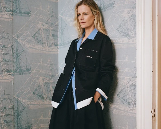 Woman in black jacket poses against wallpaper with ship design, showcasing fashion and indoor style with soft lighting.