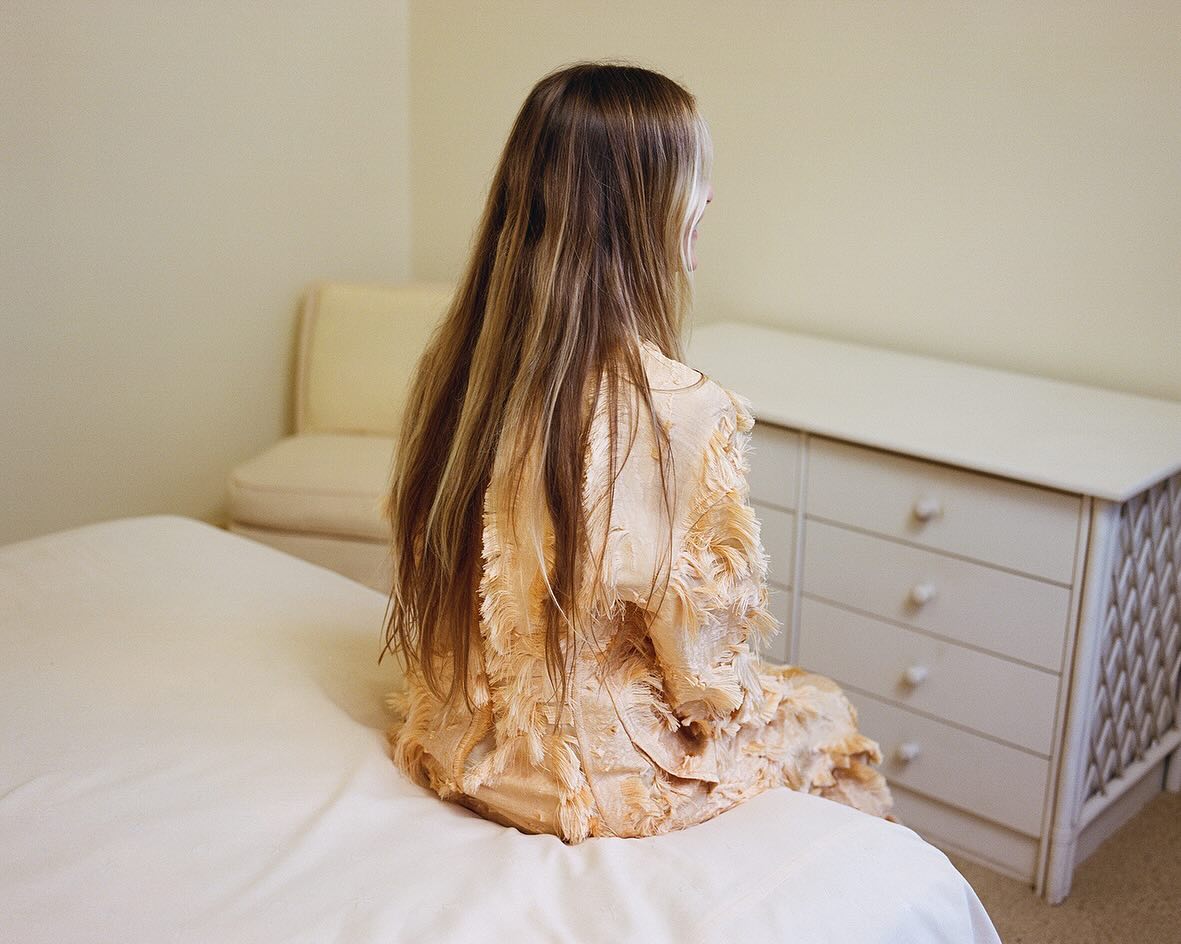 Back view of a young woman with long blond hair sitting on a bed in a bright room, wearing a light pink decorative jacket.