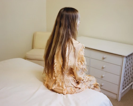 Back view of a young woman with long blond hair sitting on a bed in a bright room, wearing a light pink decorative jacket.