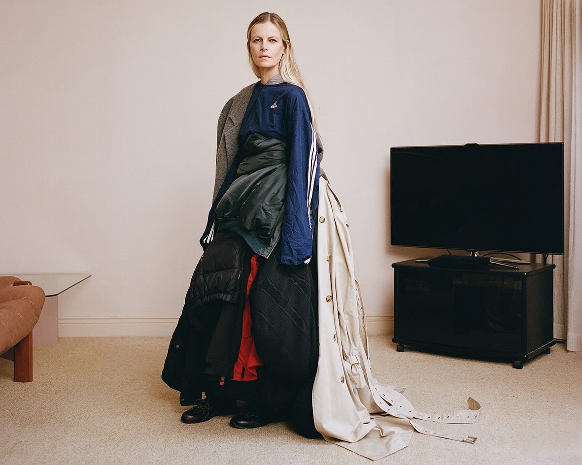 Full shot of an adult woman wearing an outfit constructed of layers of clothing standing in a room with minimal furnishings.