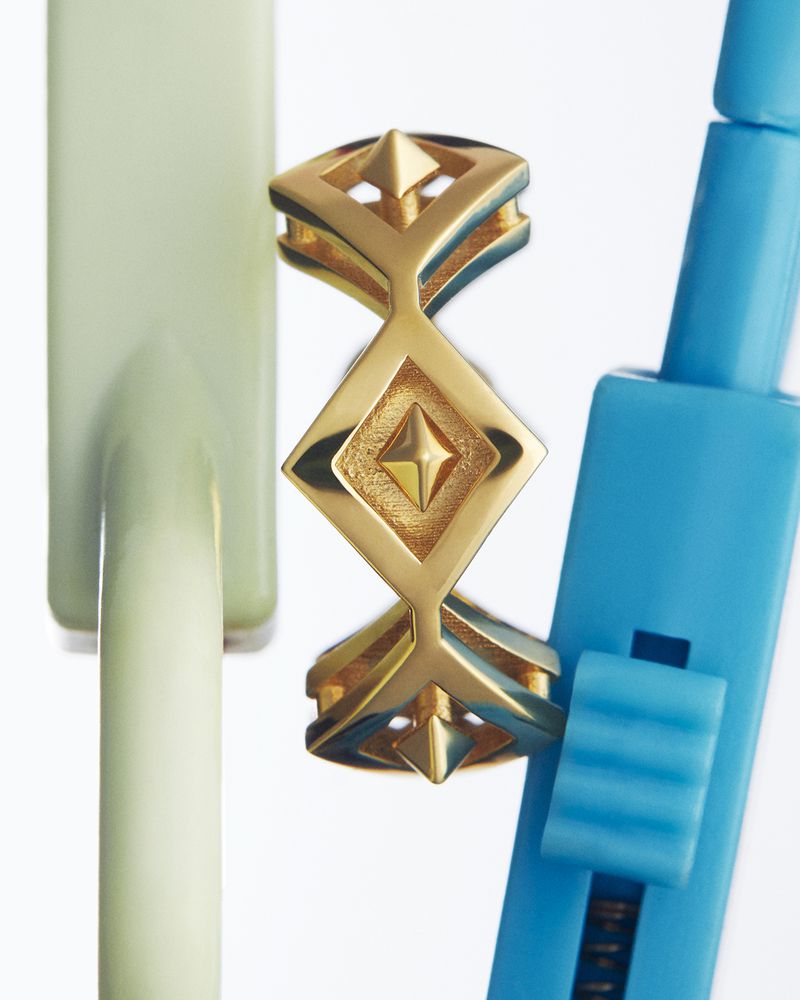 Close-up studio shot of a geometric gold ring with diamond shapes, next to blue and light green props, reflecting on a white surface.