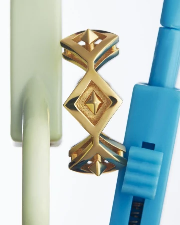 Close-up studio shot of a geometric gold ring with diamond shapes, next to blue and light green props, reflecting on a white surface.