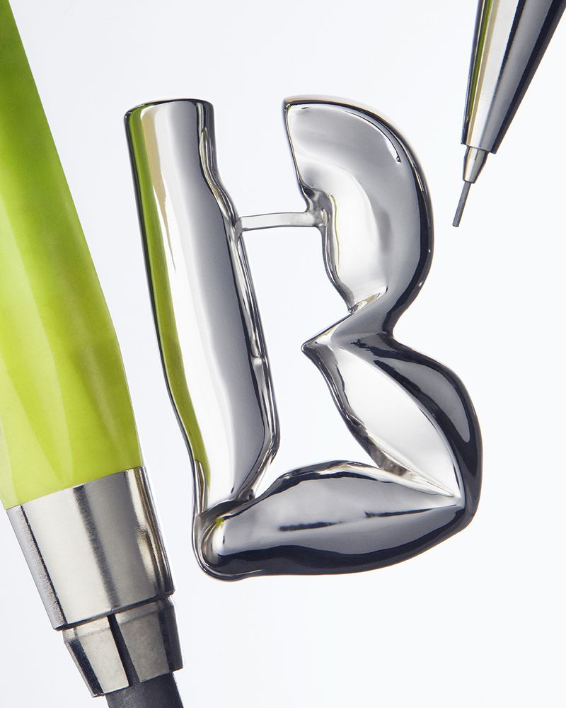Close-up studio shot: shiny, silver-colored letter B sculpture next to a green pencil and mechanical pencil against a white backdrop.