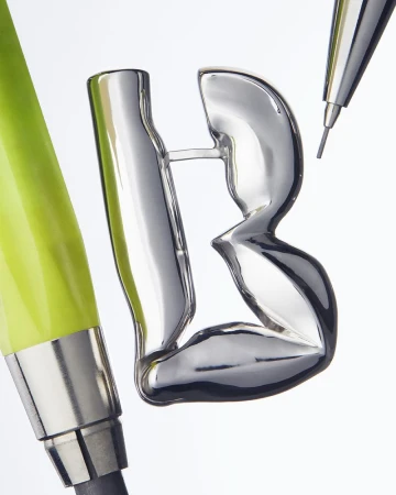 Close-up studio shot: shiny, silver-colored letter B sculpture next to a green pencil and mechanical pencil against a white backdrop.