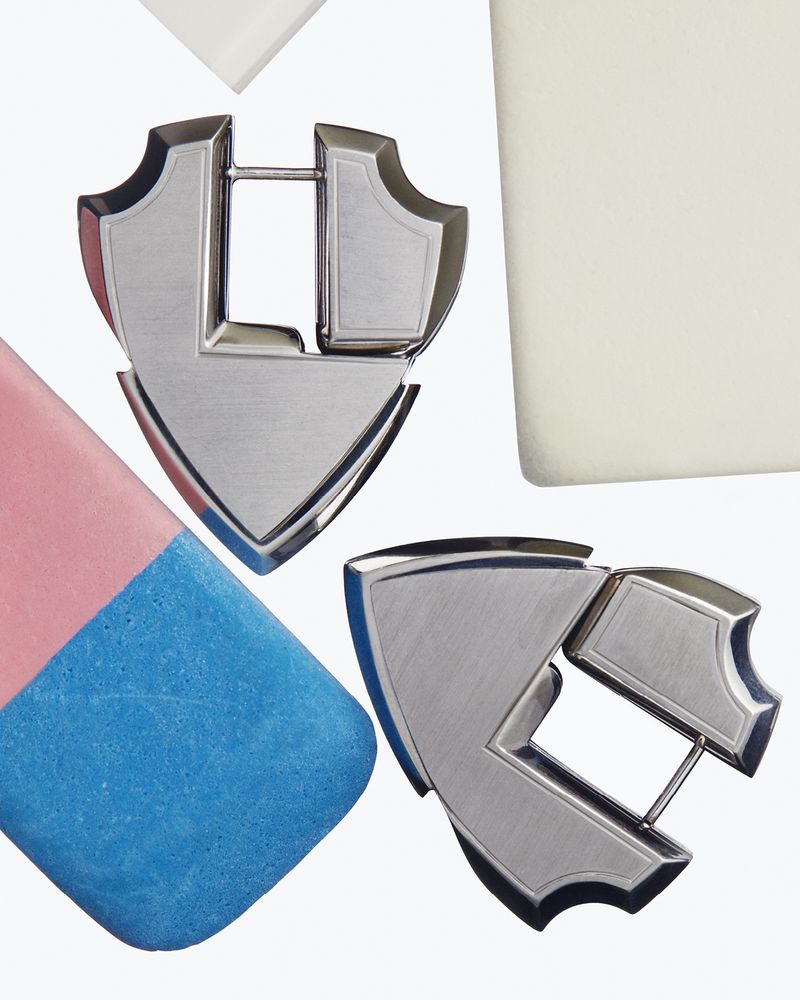 Close-up of two silver metal shield-shaped objects next to a pink and blue eraser and a white block on a white surface.