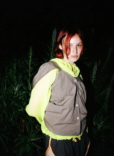 Young adult woman with auburn hair, wearing a vest and neon yellow hoodie, poses outdoors against a backdrop of dark plants.