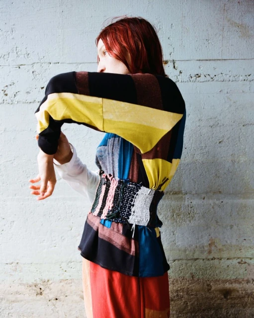 Side shot of a redhead adult female posing in a colorful jacket and orange skirt against a textured concrete wall, fashion shot.