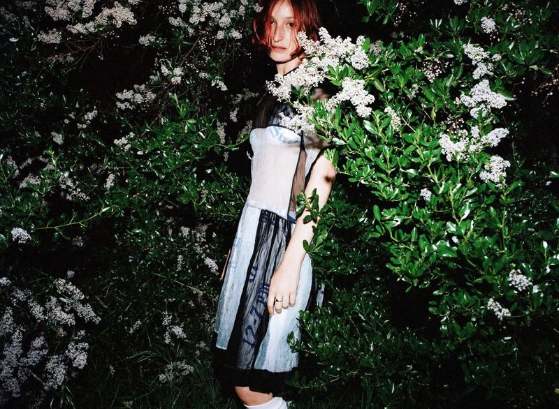 A young woman with auburn hair stands among lush green bushes with small white flowers, wearing a stylish cream and blue dress.