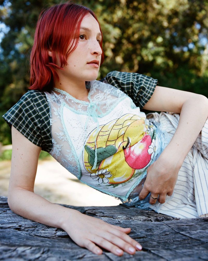 A woman with reddish-brown hair poses outdoors wearing an embellished top and striped pants, resting on a weathered wooden surface.
