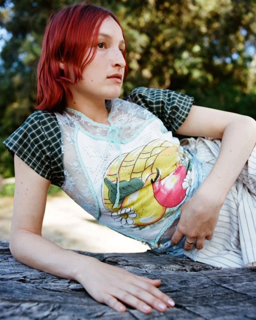 A woman with reddish-brown hair poses outdoors wearing an embellished top and striped pants, resting on a weathered wooden surface.