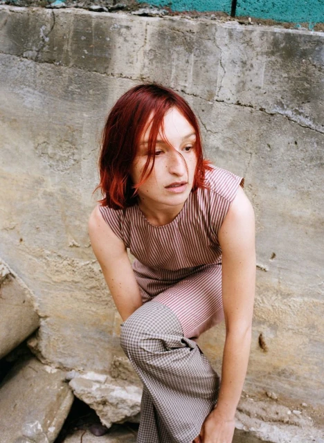 Young woman with auburn hair poses in striped top and patterned trousers in a gritty urban environment with concrete and rubble.