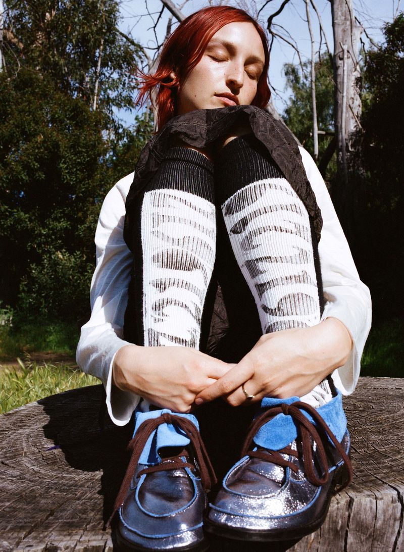Redhead woman with eyes closed sits hugging legs in patterned socks and blue shoes on wood in a forest setting.