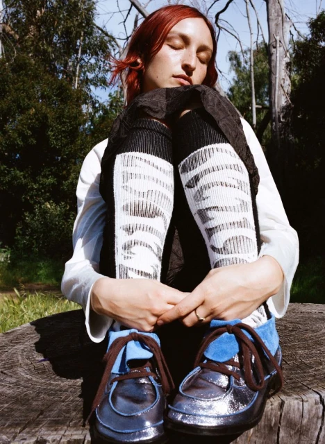 Redhead woman with eyes closed sits hugging legs in patterned socks and blue shoes on wood in a forest setting.