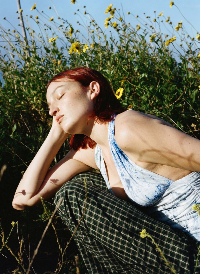 Redhead woman in patterned dress poses in sunny field of yellow wildflowers, eyes closed, resting. Outdoor fashion portrait in nature.