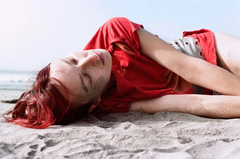 Woman with red hair lying on a sandy beach. The woman is wearing a red shirt, and the shot features close-up framing with bright lighting.