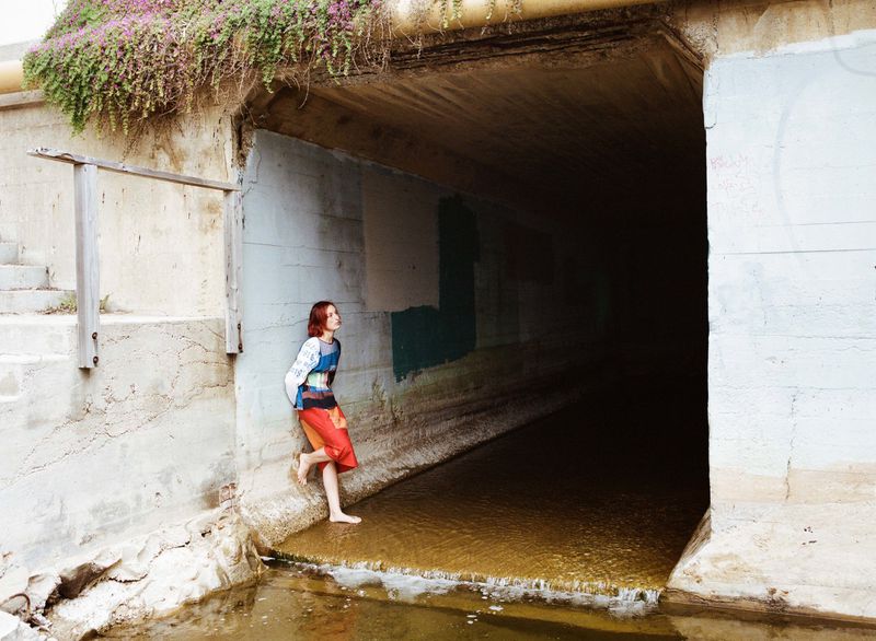 Young woman in colorful clothes stands barefoot near a concrete tunnel with water flowing through it, under an overpass covered in plants.