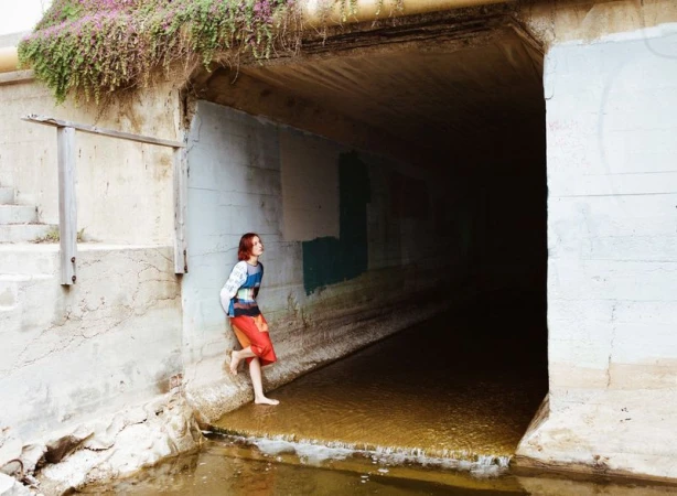 Young woman in colorful clothes stands barefoot near a concrete tunnel with water flowing through it, under an overpass covered in plants.