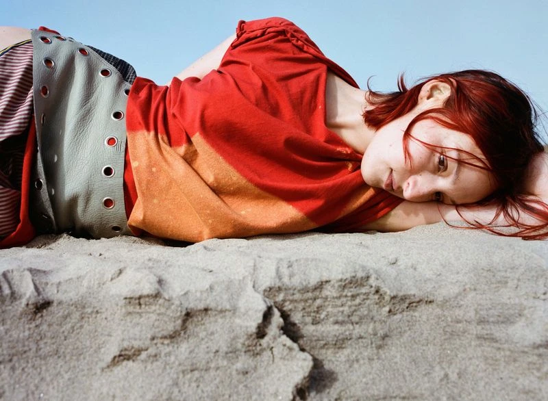 Red-haired adult woman lies on sand in orange-red shirt and patterned skirt, resting her head on her arm under a sunny sky.