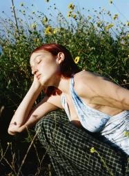 Red-haired woman with eyes closed wearing a blue patterned top and plaid trousers crouching outdoors among wildflowers