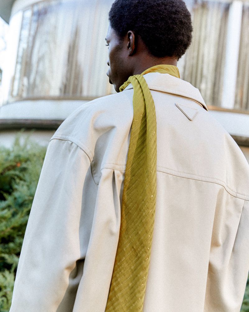 Back view of an adult Black man wearing a light beige jacket and a green patterned scarf in an outdoor setting with greenery.
