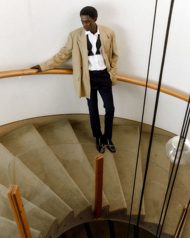 Fashionable man in suit and bow tie poses on spiral staircase in an indoor setting with neutral wall and railing in a fashion editorial.