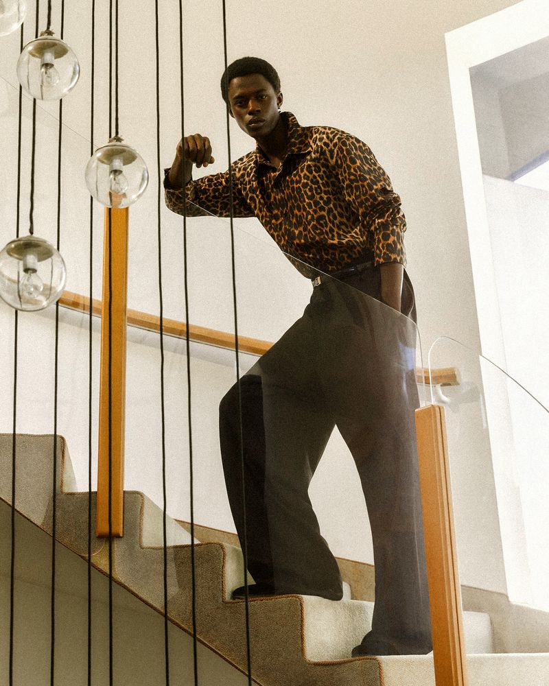 Stylish man in animal-print shirt and wide-leg pants poses on stairs with glass railing and globe string lights, fashion editorial.