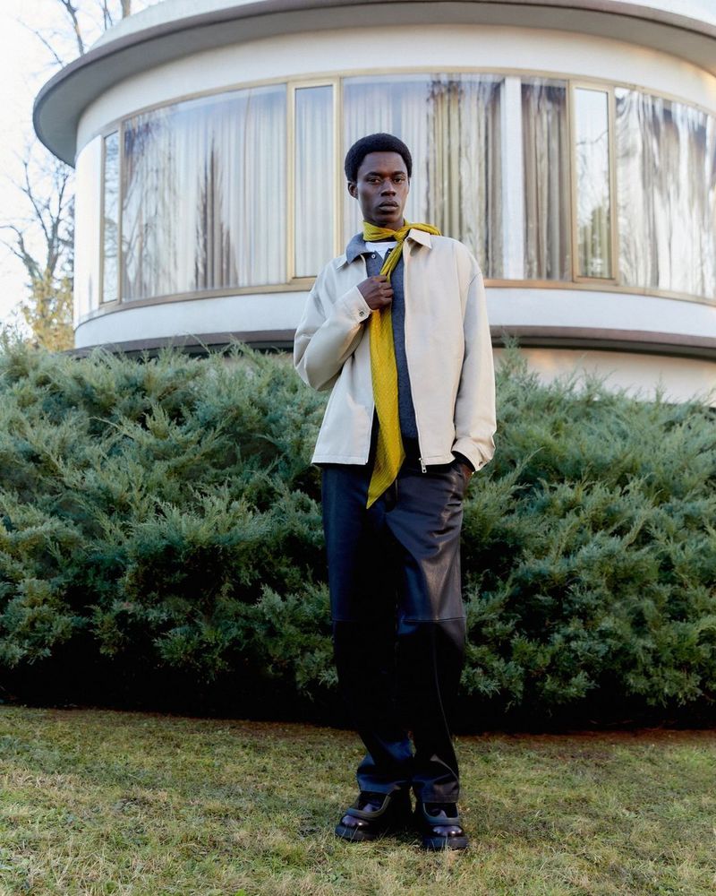 A young man standing on a lawn in a beige jacket and scarf, with a modern round house and green bushes in the background.