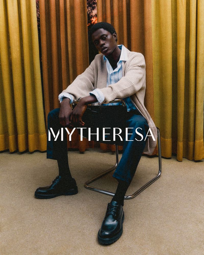 A seated African American male model wears a cardigan, light shirt, dark pants, and shoes against yellow and beige curtains.