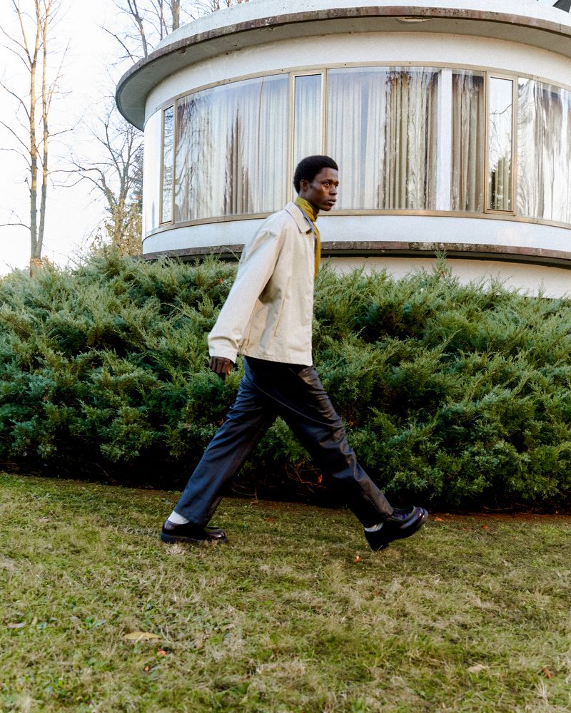 Stylish man in jacket and trousers walks on green grass in front of modern architecture. Outdoors, fashion, day.