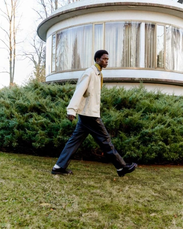 Stylish man in jacket and trousers walks on green grass in front of modern architecture. Outdoors, fashion, day.