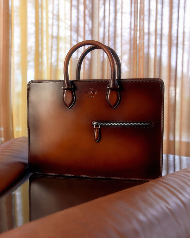A leather briefcase with a zipper pocket sits on a brown leather chair in front of neutral-toned curtains, showcasing textures and style.
