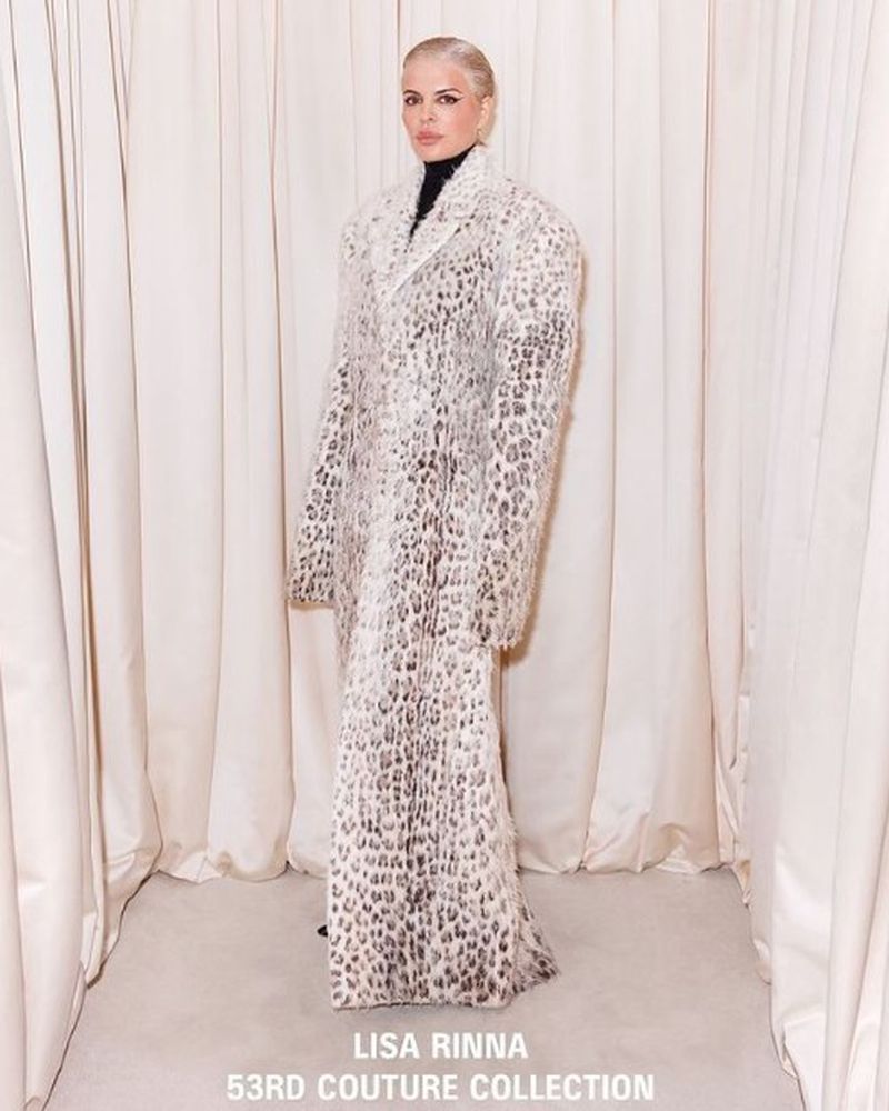 A woman poses in a full-length, long-sleeved animal print coat against a neutral curtain backdrop for a fashion editorial photo.