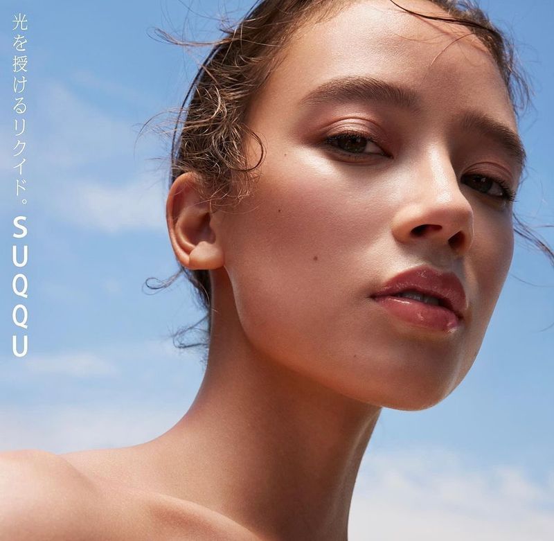 Close-up portrait of a young woman with light skin and wet hair against a bright blue sky, featuring Japanese brand logo and beauty advertising.
