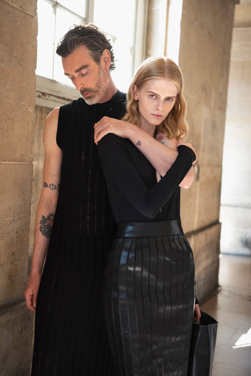A light-skinned man and woman embrace in a fashion shot; the man wears a black tunic, and the woman wears a black top and leather skirt.