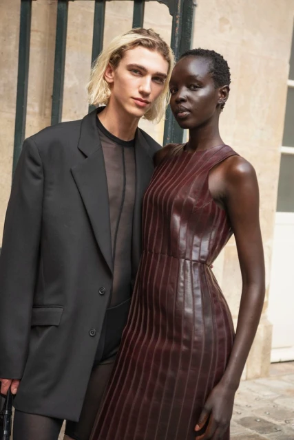 Two fashion models pose outdoors. One is a blonde male in black mesh top and suit, the other a Black female in a burgundy dress.