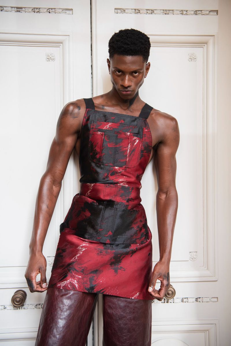 Portrait of a Black male fashion model wearing a red and black outfit, standing against a white door with a serious facial expression.