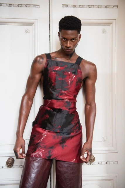 Portrait of a Black male fashion model wearing a red and black outfit, standing against a white door with a serious facial expression.
