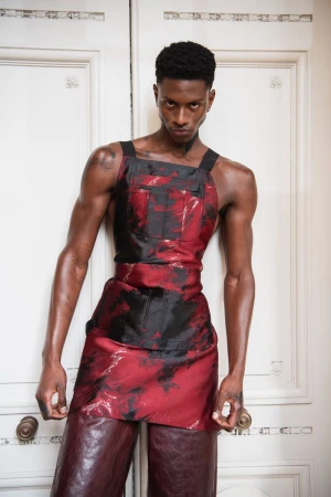 Portrait of a Black male fashion model wearing a red and black outfit, standing against a white door with a serious facial expression.