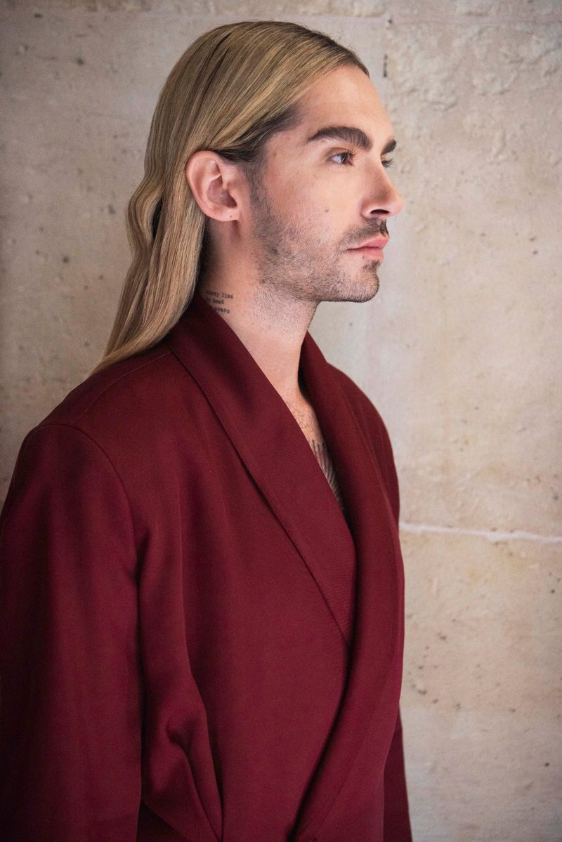 Profile shot of a man with long blonde hair and a mustache wearing a maroon robe standing in front of a neutral colored wall.
