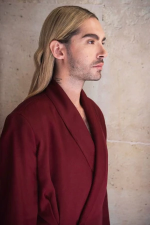 Profile shot of a man with long blonde hair and a mustache wearing a maroon robe standing in front of a neutral colored wall.