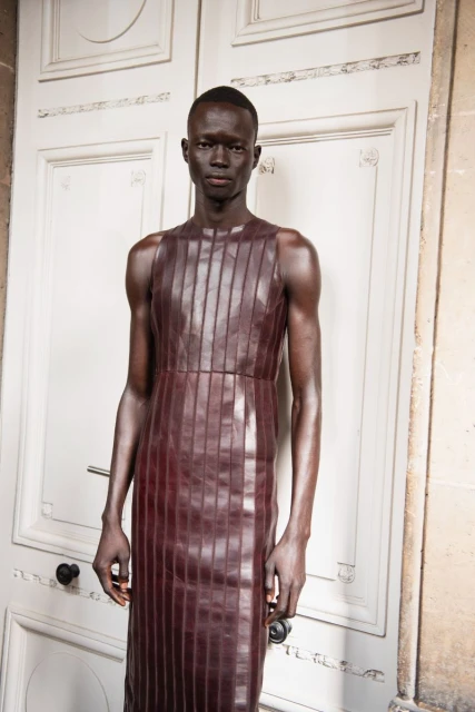 Portrait of a Black male model in a brown leather dress against a white door, showcasing fashion and style in an indoor setting.