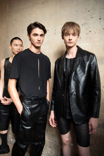 Three adult males in black attire pose against a neutral wall. The two men in front wear a t-shirt and leather jacket, respectively.
