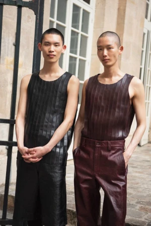 Two slender Asian men pose side-by-side outdoors. Both wear dark leather-look tops with vertical striped details and matching pants.