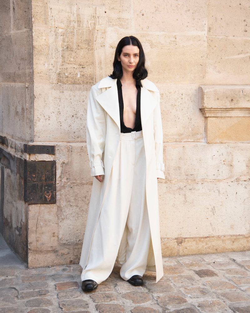Woman in oversized white coat and wide-leg trousers stands in an urban setting with a stone wall, showcasing a fashionable, relaxed style.