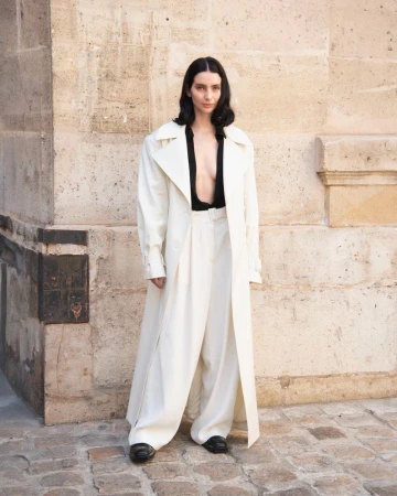Woman in oversized white coat and wide-leg trousers stands in an urban setting with a stone wall, showcasing a fashionable, relaxed style.