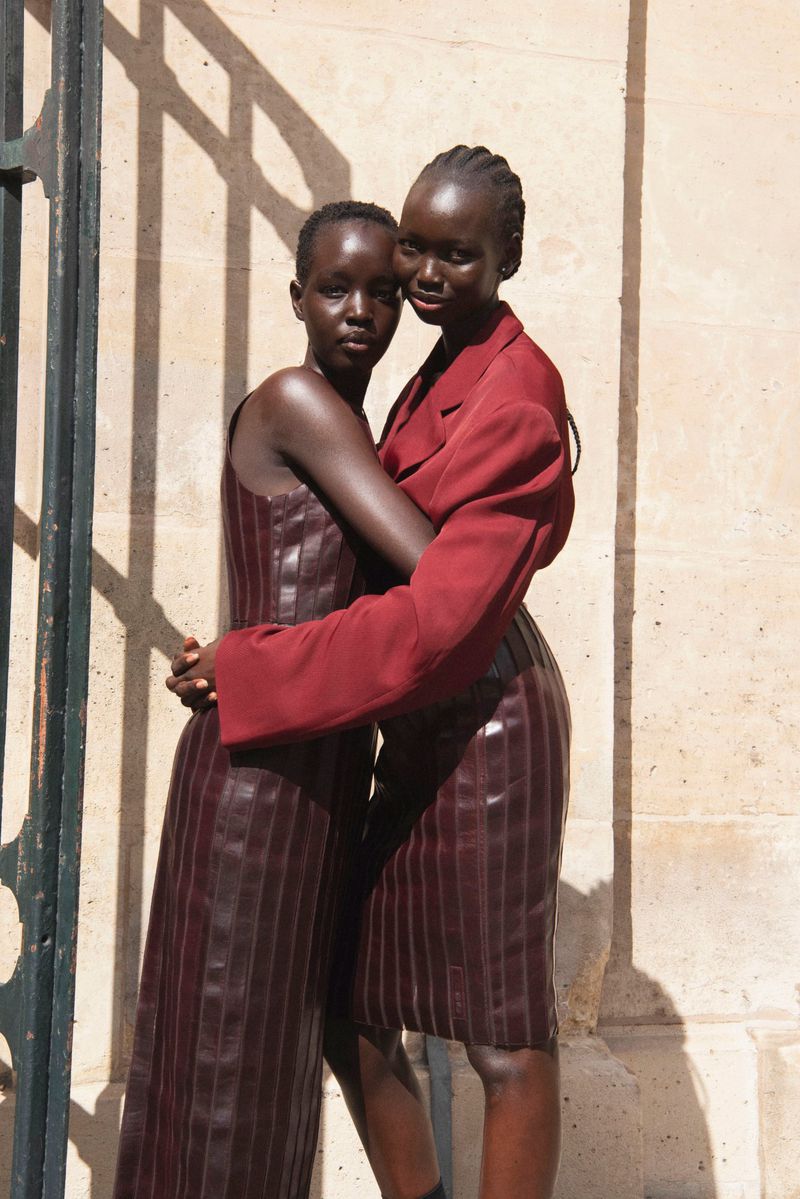Two women embrace in a fashion shot. One wears a dark red dress, and the other wears a jacket and skirt. They stand in front of a stone wall.