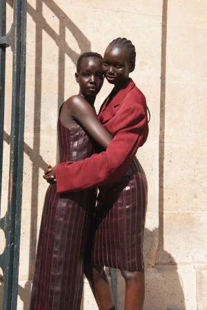Two women embrace in a fashion shot. One wears a dark red dress, and the other wears a jacket and skirt. They stand in front of a stone wall.