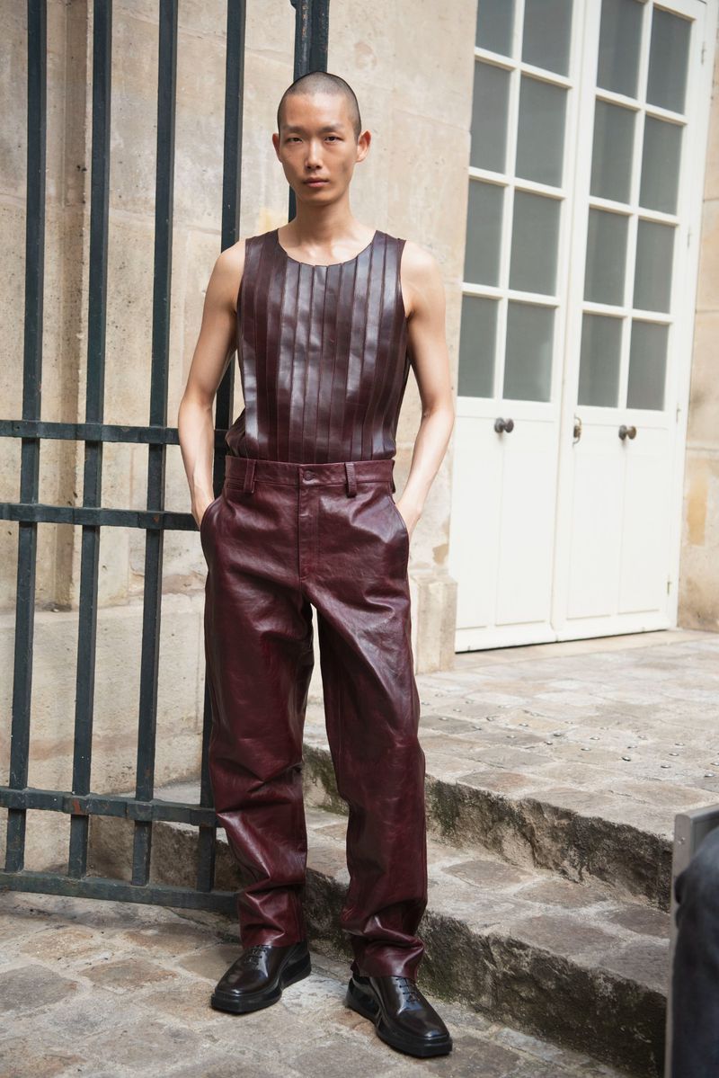 Full shot of a bald, Asian male model in burgundy leather top and trousers, posing in front of a beige stone building and black iron fence.