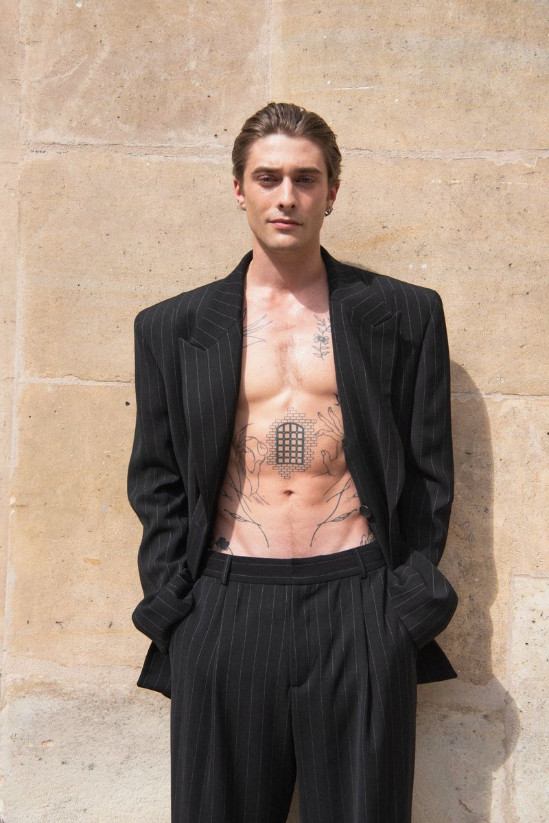 Adult male poses outdoors wearing a pinstripe blazer and matching pinstripe pants with no shirt showing tattoos on his chest and stomach.