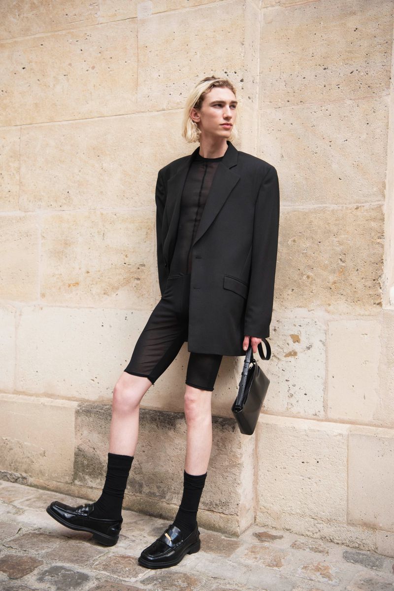 A slender adult male with platinum-blonde hair in a black blazer, sheer black shirt, and black shorts poses in front of a tan stone wall.